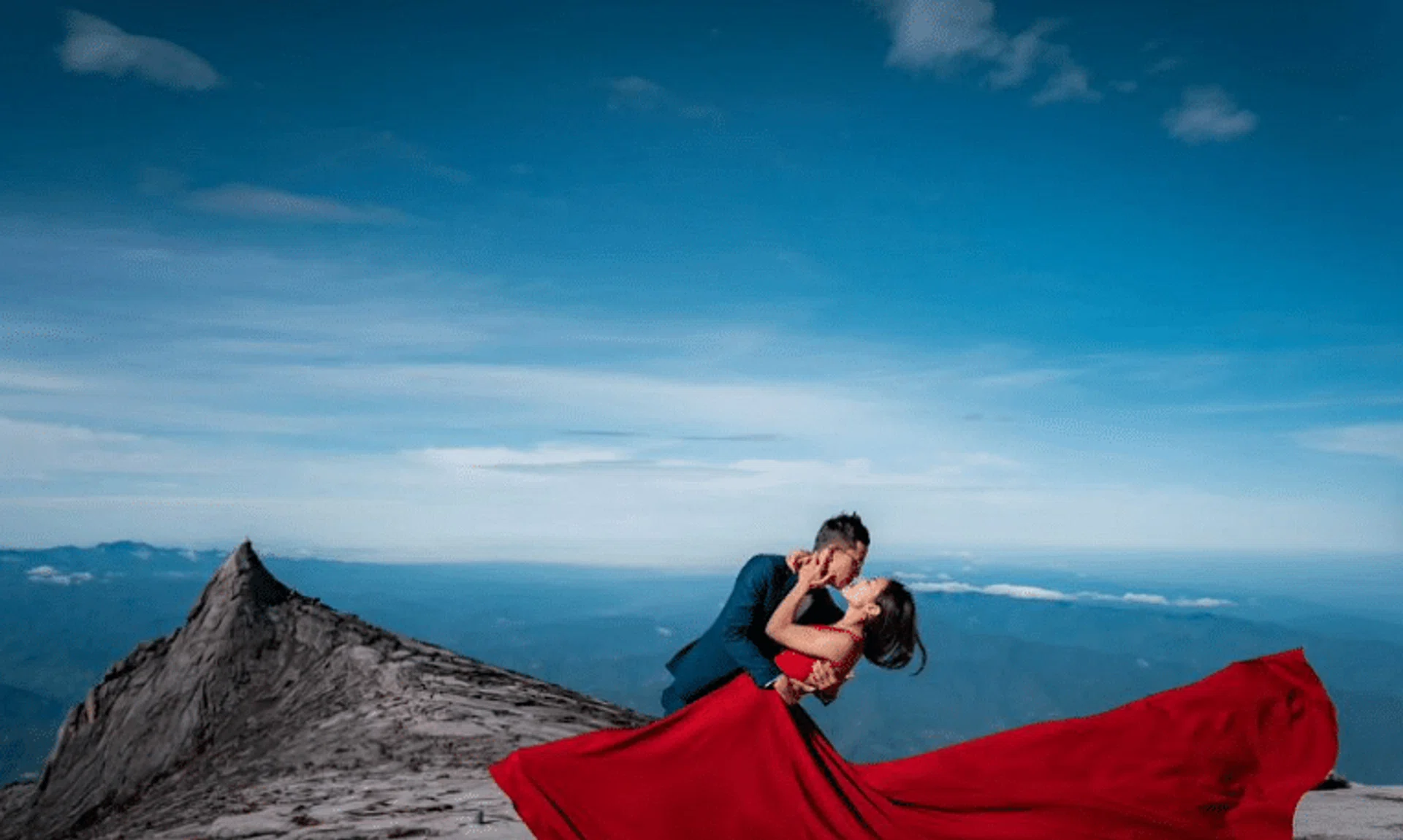 The bride-to-be, dressed in a red sleeveless dress, and her partner, clad in a navy suit with a bowtie, pose with South Peak in the background.