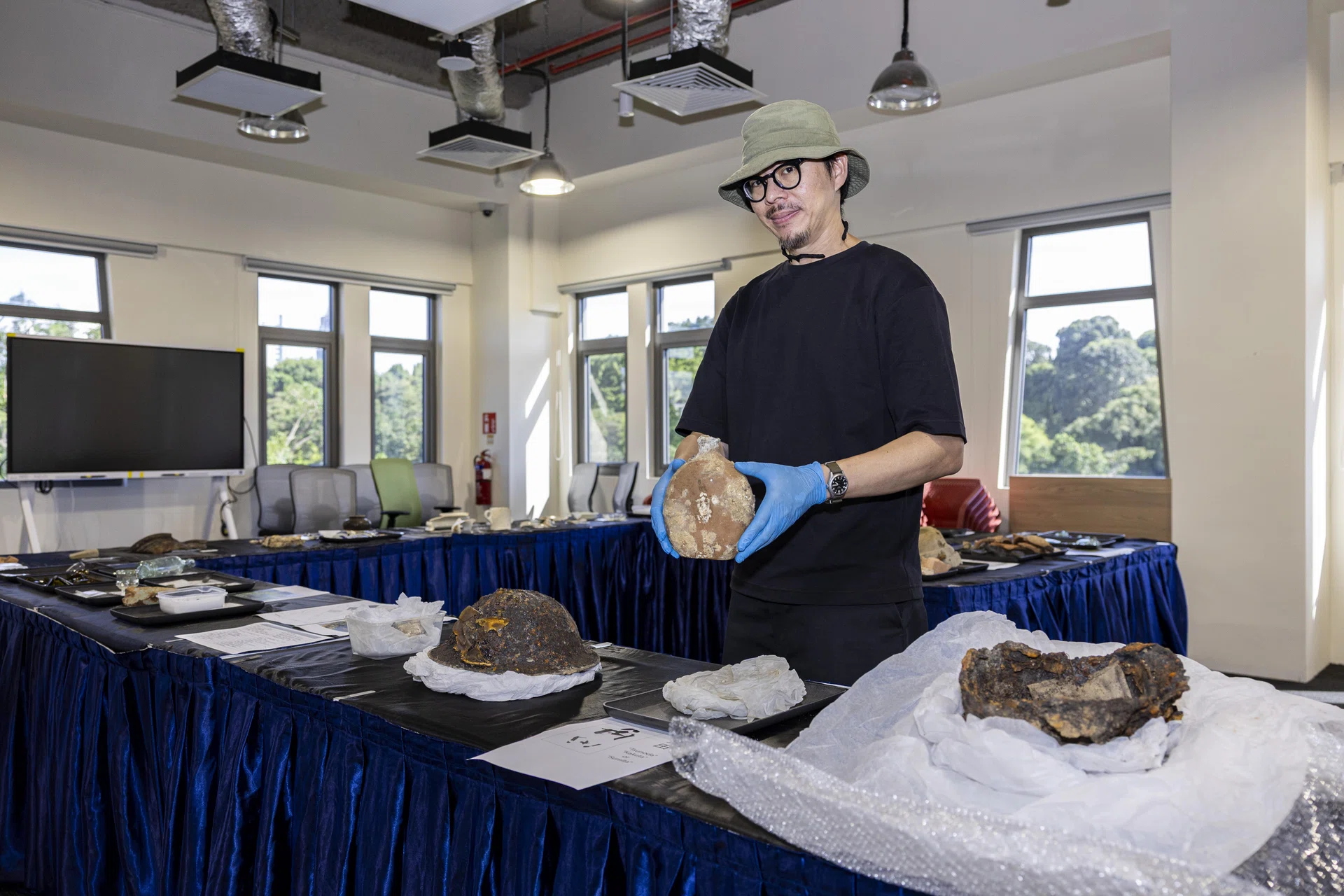 Archaeologist Aaron Kao holding a Japanese army water bottle that was found in Alexandra Hospital. On the table are helmets that were also recovered from the hospital's grounds.