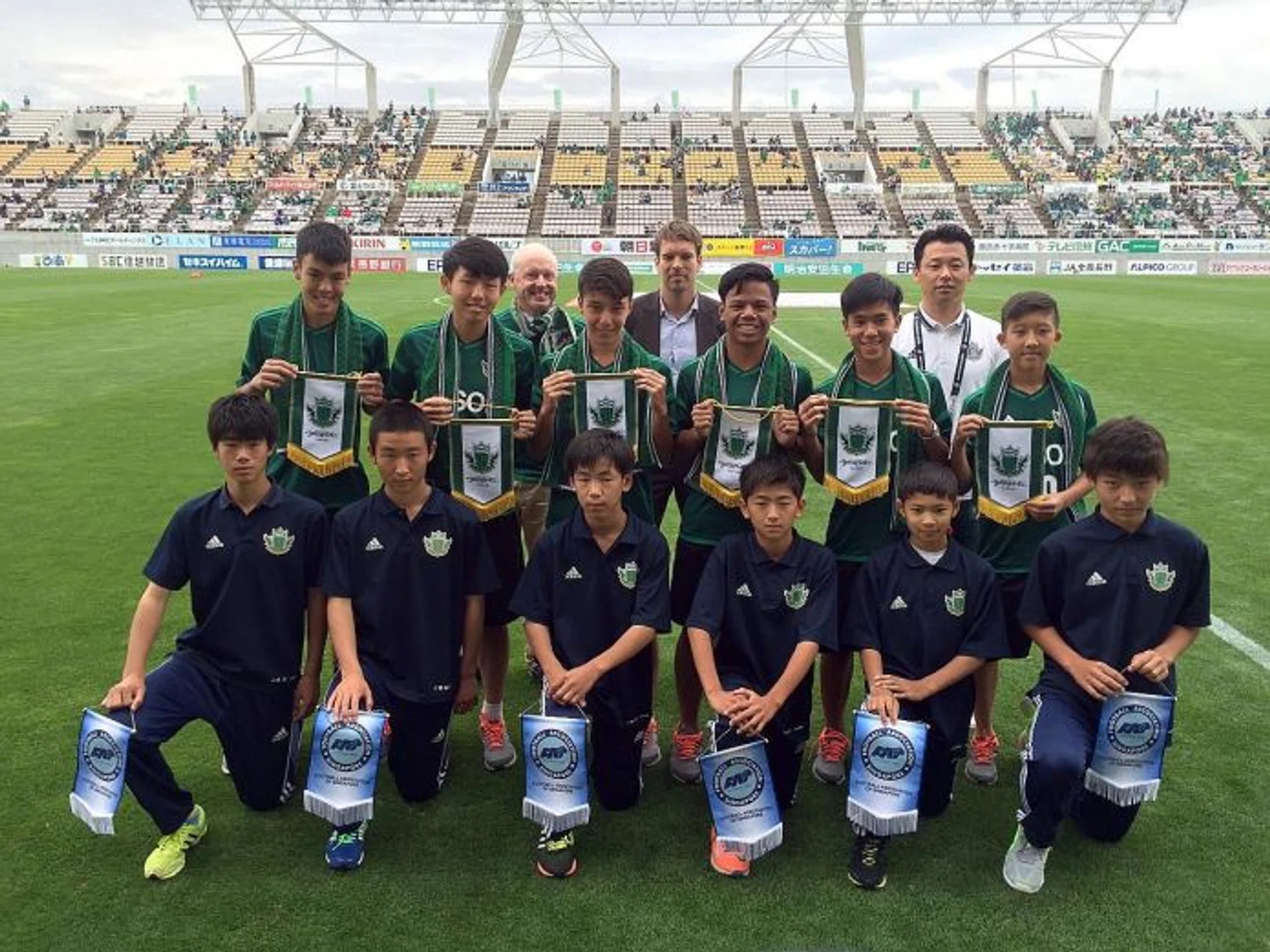 ENRICHING EXPERIENCE: NFA boys (from left, in green) Vasileios Chua, Ashley Yong, Christian Chiang Moroni, Nur Adam Abdullah, Marc Tan and Elijah Lim exchanging pennants with their Matsumoto Yamaga counterparts (front row) at the Alwin Stadium before Yamaga beat Montedio Yamagata 1-0 in a J2 League match.
