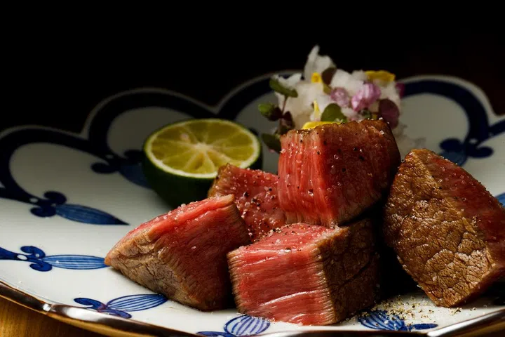 Five cubes of kobe wagyu, grilled to rare, are presented on a plate with garnish on top of the meat and lime to the side.