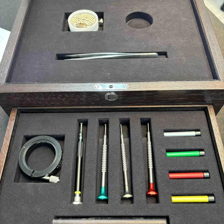 An open Chopard watchmaking training toolkit displayed in a fitted wooden presentation case. The set contains anti-magnetic tweezers, a watchmaker’s loupe, four precision screwdrivers with colour-coded handles and additional interchangeable screwdriver grips, each arranged in custom-cut foam compartments.