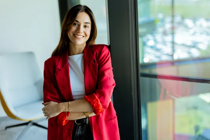 Young woman stands confidently with a smile, dressed in a stylish red blazer, leaning against a glass window in a contemporary office space