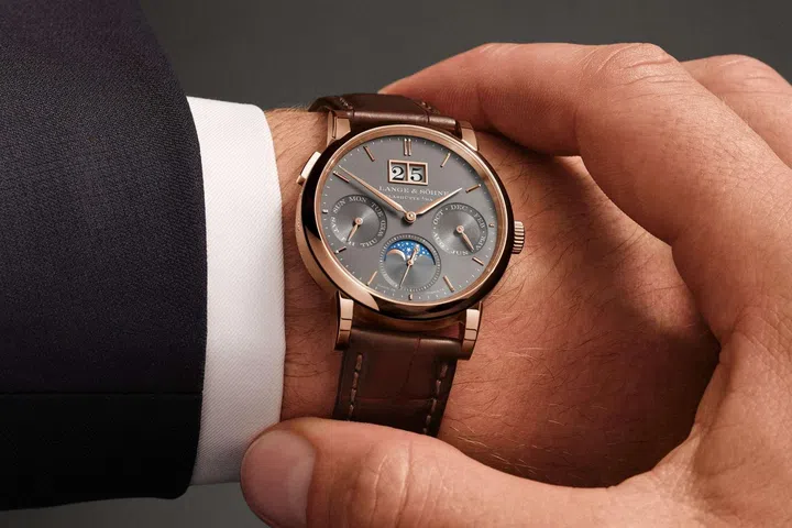 The A. Lange & Söhne Saxonia Annual Calendar in pink gold with a grey dial, displaying the outsize date at 12 o’clock, day and month subdials, subsidiary seconds, and a blue moon-phase display within a refined 36mm case.