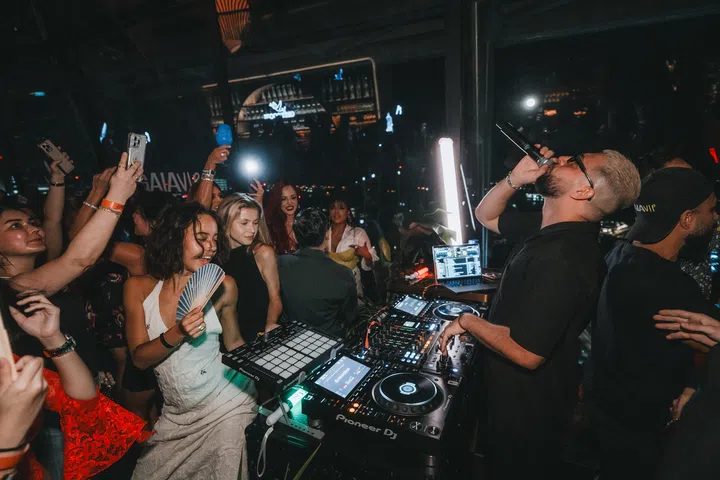 A man stands behind a DJ booth, speaking into a microphone, while a crowd dances in front of him, the first few clearly distinct and visible