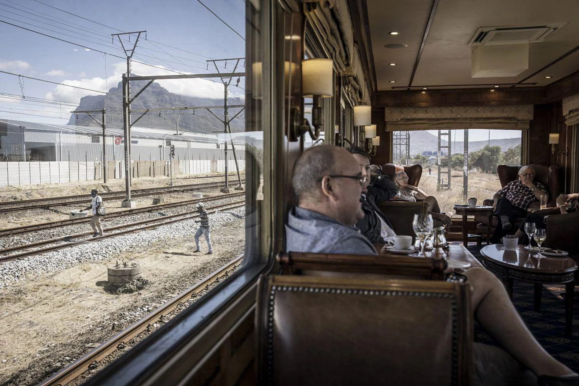 Blue Train passengers enjoy drinks in the observation car as the train slowly makes its way through the outskirts of Cape Town, on February 25, 2021. - Forty-eight hours to stop thinking about anything, to let yourself be pampered, to play the princes and princesses. The Blue Train, the ultimate luxury travel train experience,  crosses South Africa for more than 1,400 km from Cape Town, its southern tip, to its capital Pretoria, through fields and slums, urban centres and dream landscapes. (Photo by MARCO LONGARI / AFP)
