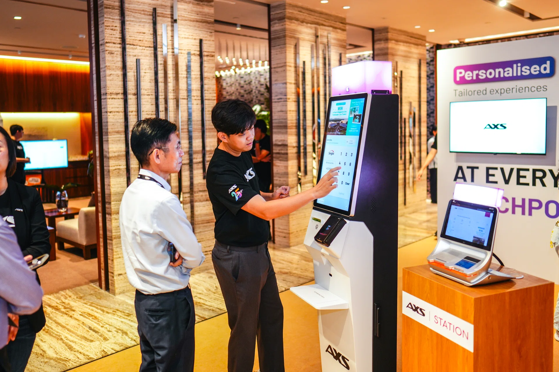 A man stands in front of a digital kiosk, showing another man how to use it.
