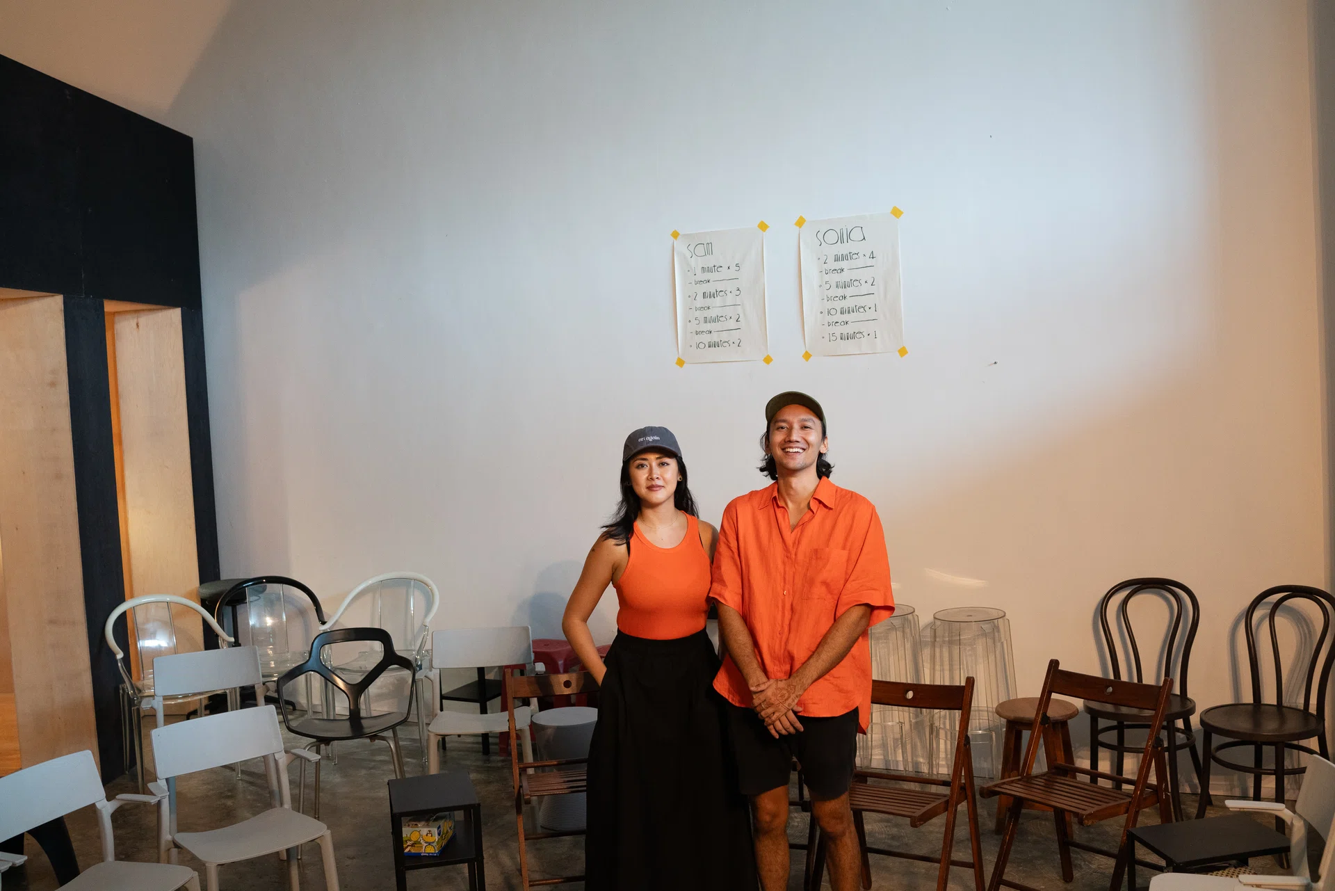 Two individuals wearing orange and caps, on the left is a woman , co-founder Chingyi Chua, while on the right is a man, co-founder Milon goh. Both stand in the center of a room with chairs