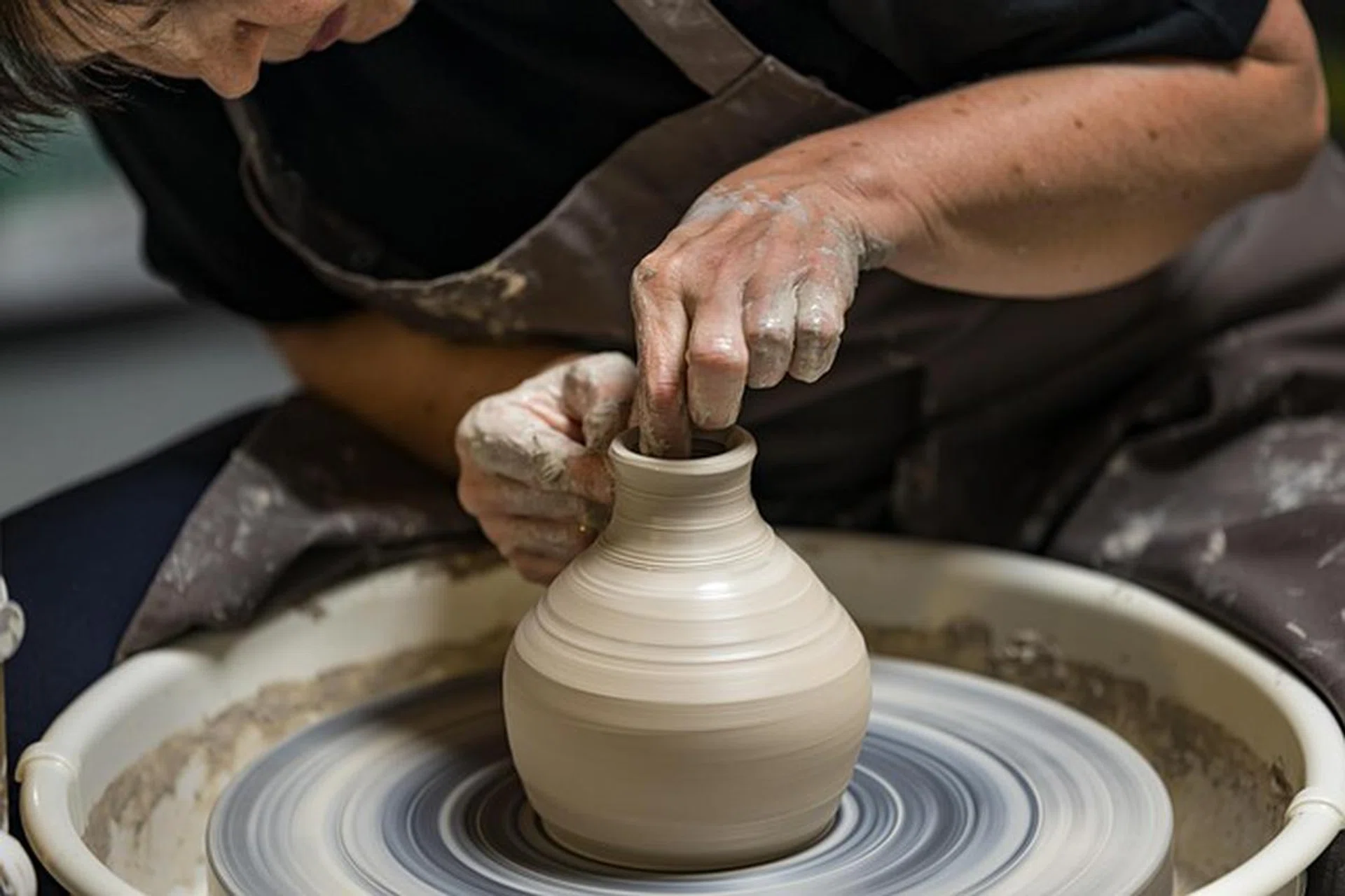 A pottery wheel, with a person behind, leaning over and shaping clay into a pot. The pot has a wide bottom and small mouth.