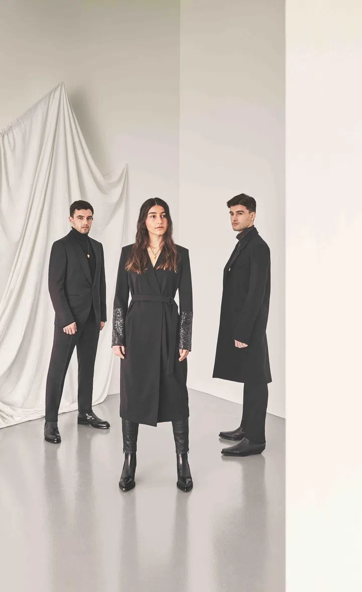 Carmen is wearing a black wool long coat with sequinned sleeve detail and black leather tights from Brunello Cucinelli paired with black leather boots from Bottega Veneta. Gabriel is wearing a black wool turtleneck, black silk tuxedo suit and black leather boots from Ermenegildo Zegna. Rogerio is wearing a dark brown viscose turtleneck, black wool long coat, black wool trousers, black leather lean boots from Bottega Veneta.
