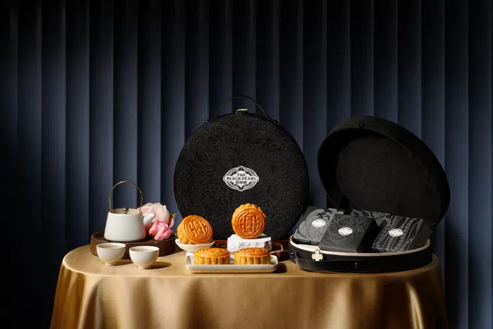 Four mooncakes arranged at the center of a table with a gold tablecloth, with two circle shaped boxes, one closed and stood up behind the mooncakes, the other opened on the mooncakes’ right. On the left of the table is a teapot and two tea cups.