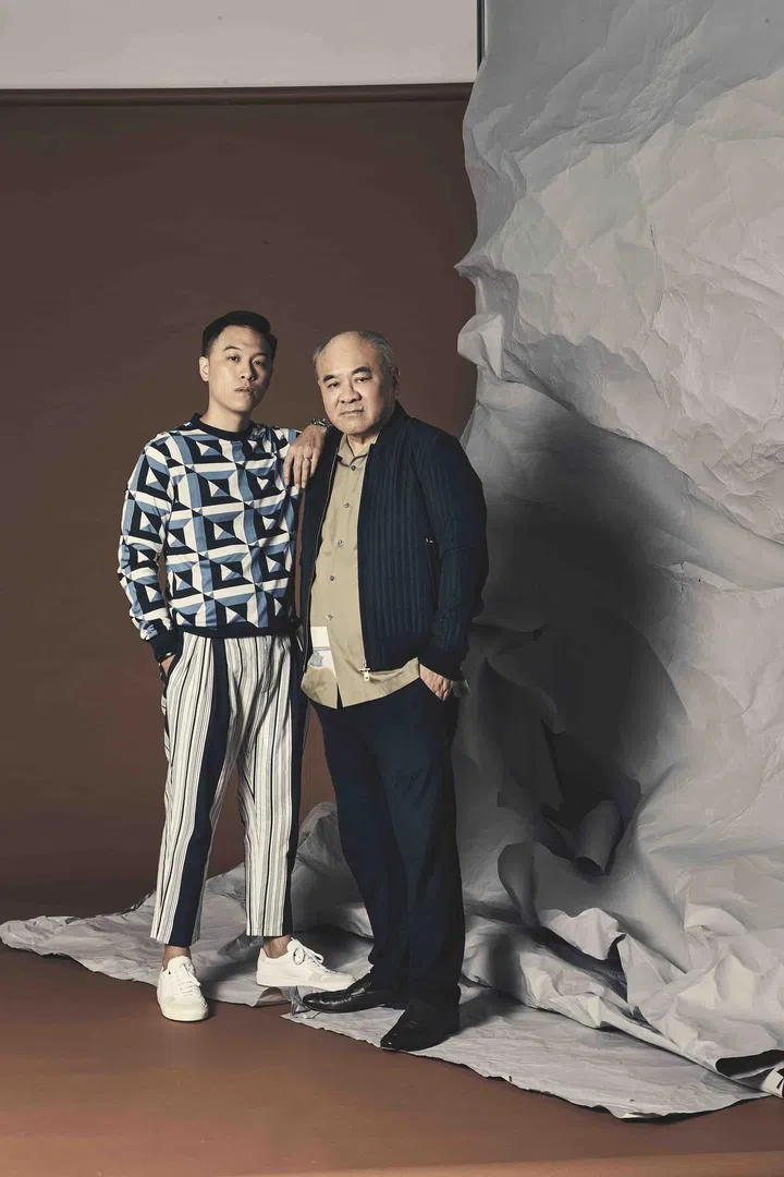 Jon is wearing a geometric print cotton knit sweater andcotton striped trousers from Dolce & Gabbana, and white leather sneakers, from Boss. Jon's father is wearing a cotton short sleeved shirt and black quilted bomber jacket from Dunhill.