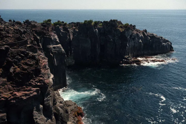 Rugged black volcanic cliffs at Jogasaki Coast on Japan’s Izu Peninsula, where foaming Pacific waves crash against lava-formed rock formations beneath a clear blue sky, showcasing the dramatic coastal landscape that inspired Grand Seiko’s Ushio collection.