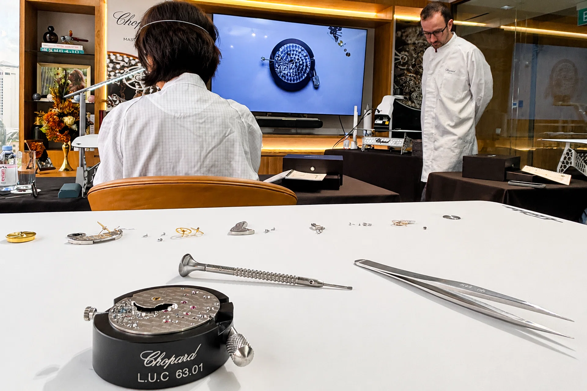 A Chopard watchmaking classroom with participants seated at individual workbenches as an instructor demonstrates techniques at the front. In the foreground, a decorated L.U.C Calibre 63.03-C training movement rests on a work stand alongside precision tools.