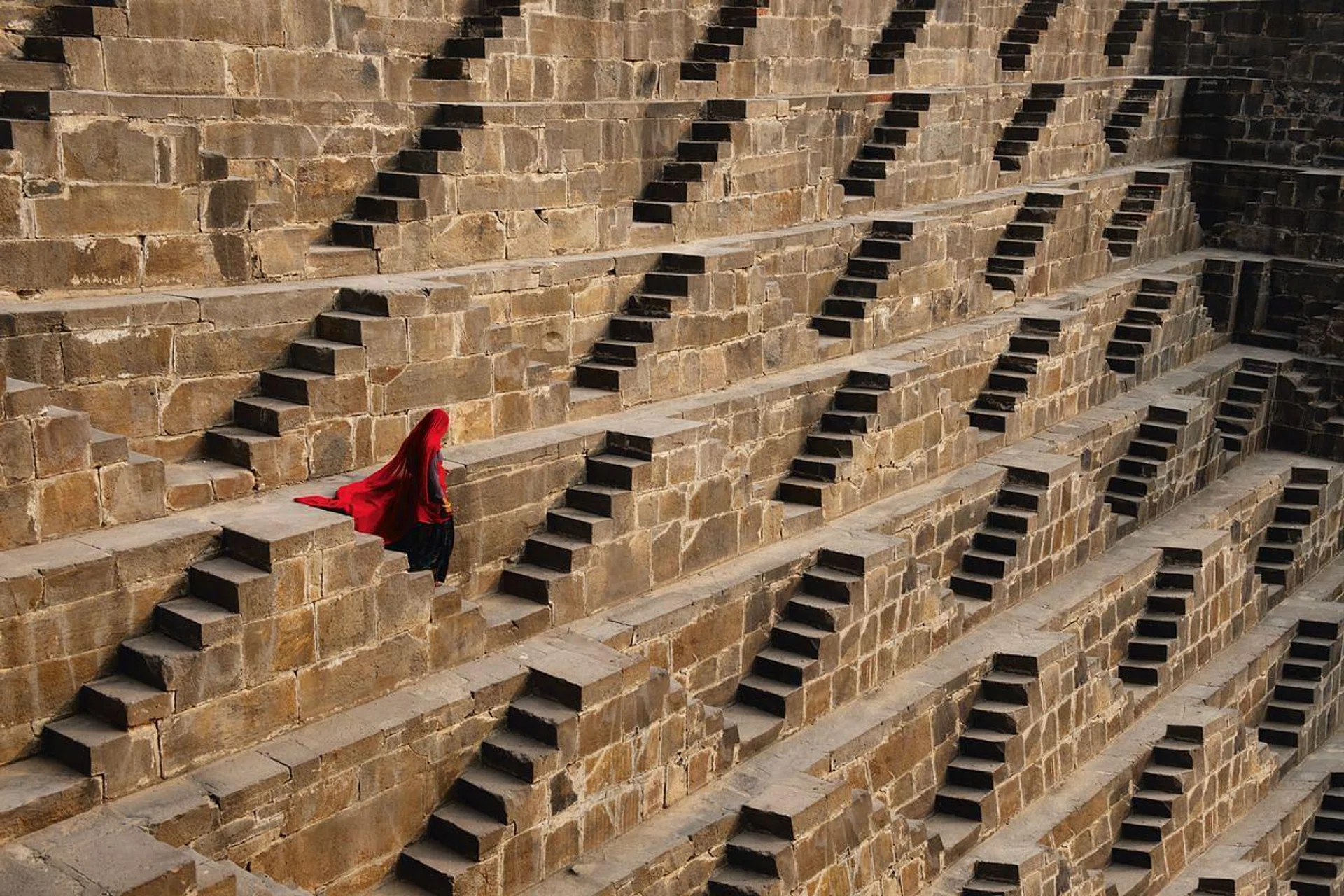 <B>A Time for Contemplation</b><br>
CHAND BAORI STEPWELL, ABHANERI, RAJASTHAN, INDIA<br><br>
Located near a temple in Abhaneri, Chand Baori Stepwell is a historical centre of social life for the locals, who meditatively descend the myriad steps to reach the water. 
<hr><i>
“The place immediately reminded me of the graphic work of the artist Maurits Cornelis Escher, notably expressed through his art of creating optical illusions through playing with lines. The Indian women we met appeared to be lost amid the vastness of the monument. These are definitely the most beautiful and awe-inspiring staircases I have ever seen. The architects worked miracles in creating a work of pure beauty.”</i>