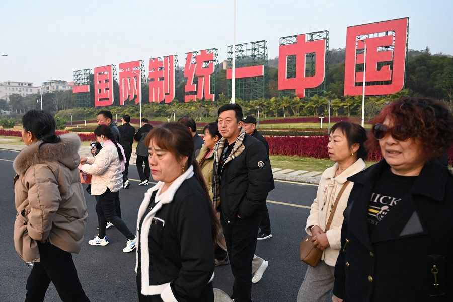 People walk past a propaganda slogan which reads "One Country, Two Systems, Reunify China", which can be seen from Taiwan's Kinmen island, on a road at the waterfront in Xiamen, Fujian province, China, on 12 January 2024, the day before Taiwan's presidential election. (Greg Baker/AFP)