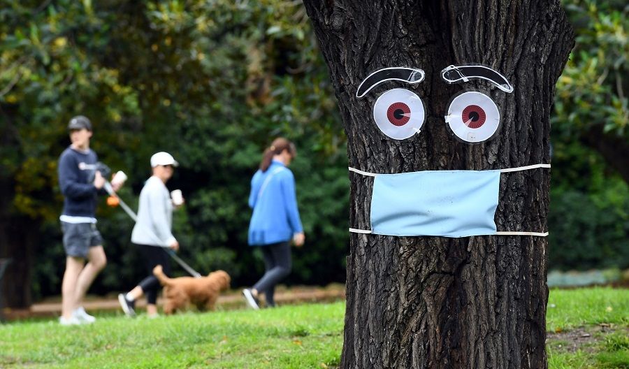 People walk past a tree with a mask and eyes stapled on it, in Melbourne, on 20 April 2020. (William West/AFP)