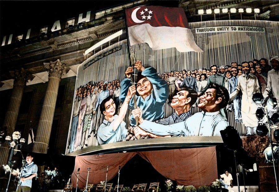 Prime Minister Lee Kuan Yew goes through an overnight rehearsal of a ceremony at City Hall on the eve of Singapore's first anniversary in 1966. A large image shows Singapore's various ethnic groups holding up the national flag, symbolising the nation coming together to forge a bright future.
