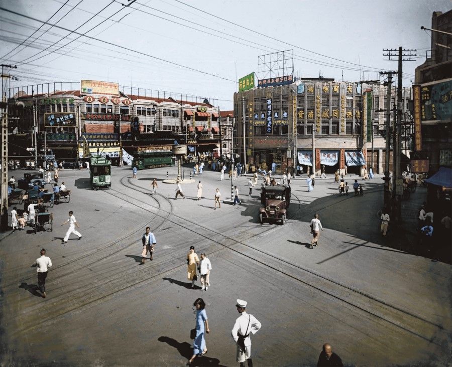 A view of the entrance to old Ximen Street in Shanghai's old city during the Republic. People traverse the wide street in twos and threes, as rickshaws, cars, and trams vie for the right of way, with plenty of stores and signs along the street.