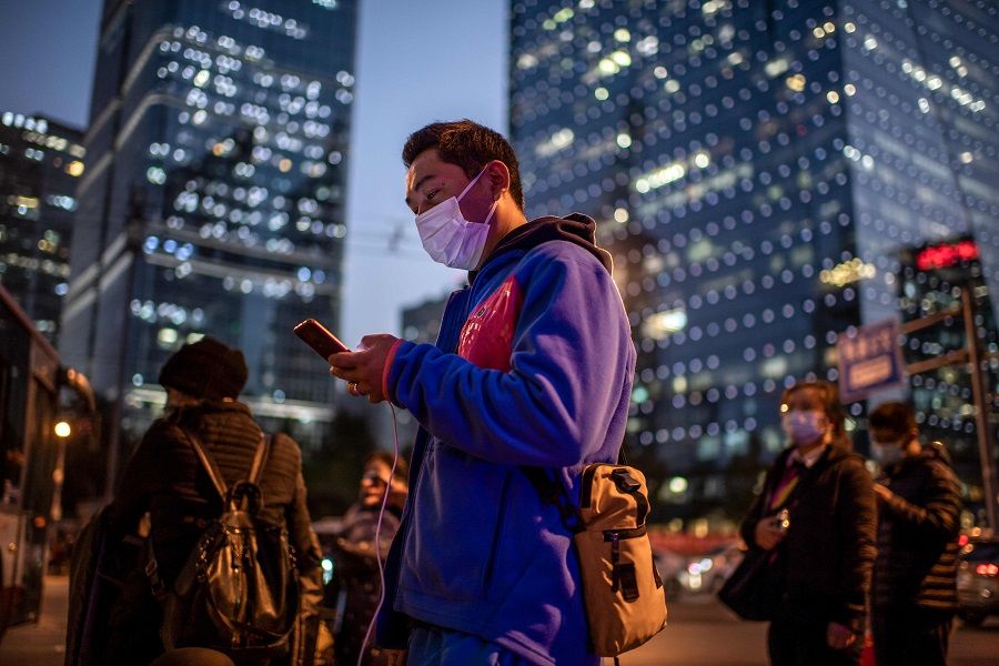 A man (centre) wearing a face mask looks at his mobile phone as he waits to cross a street during rush hour in Beijing on 21 October 2020. (Nicolas Asfouri/AFP)