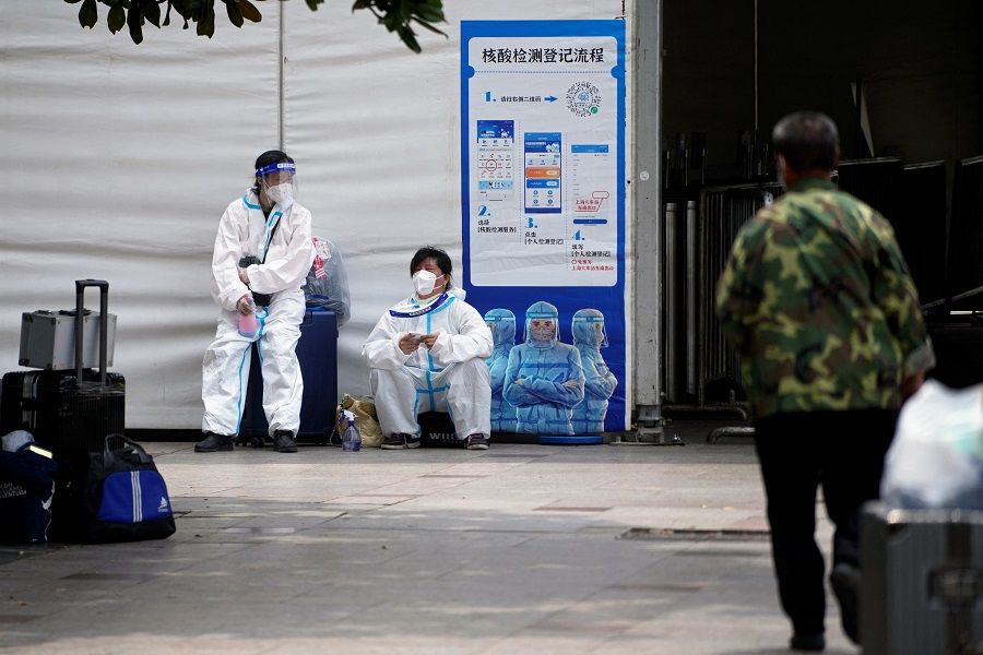 People in protective suits sit on suitcases near a sign for PCR testing registration outside the Shanghai Railway Station, China, 1 June 2022. (Aly Song/Reuters)