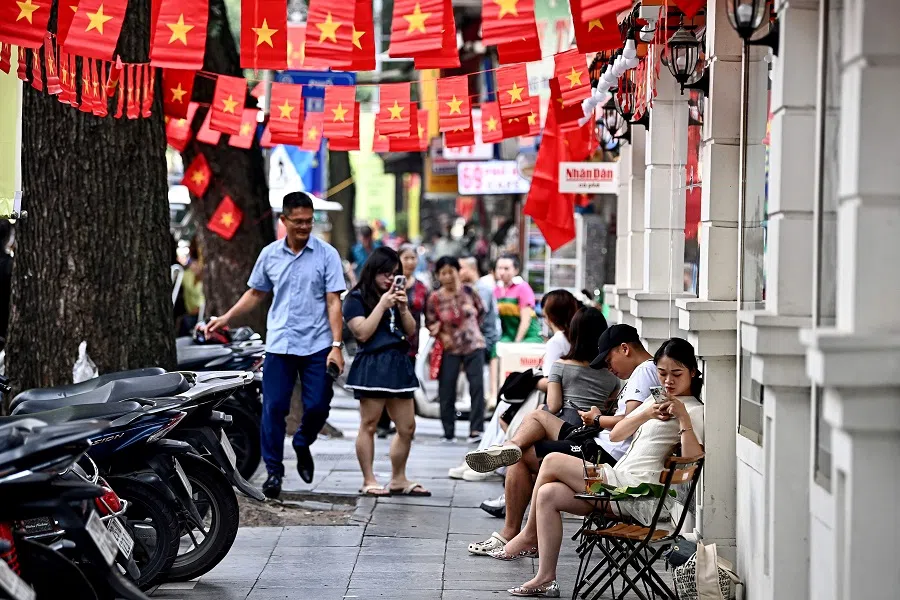 People sit and drink coffee next to Vietnamese flags displayed above a sidewalk in Hanoi on 28 August 2024.