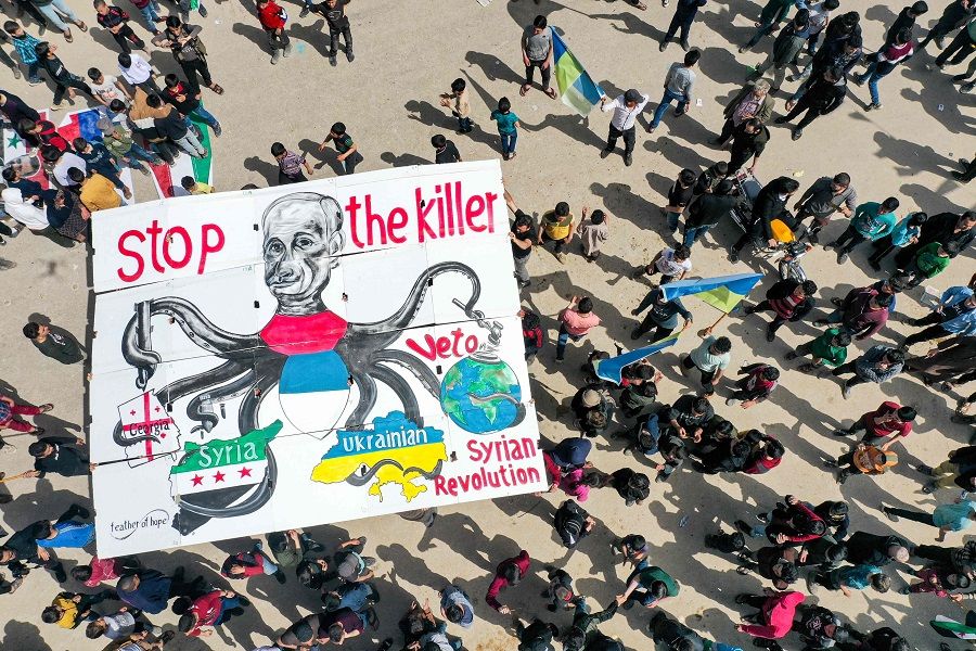This picture taken on 1 April 2022 shows an aerial view of a giant sign being raised by protesters depicting Russia's President Vladimir Putin as an octopus with its arms wrapping around the countries of Georgia, Syria, Ukraine and the world globe during a demonstration in the city of Binnish in Syria's northwestern rebel-held Idlib province against Russia's invasion of Ukraine. (Omar Haj Kadour/AFP)
