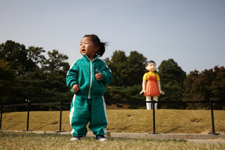 A girl wearing a costume of Netflix series Squid Game poses for photographs in front of a giant doll named 'Younghee' from the series on display at a park in Seoul, South Korea, 26 October 2021. (Kim Hong-Ji/Reuters)