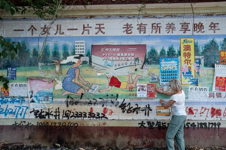 The photo taken on 6 June 2024 shows Corinne Wilson sticking a flyer in the hope of finding her adopted daughter Loulee’s biological parents on a faded billboard with a slogan that reads “A daughter is like a slice of heaven, she’ll take good care of you when you’re old”, which was used to promote China’s “one-child” policy in Dianjiang county, Chongqing municipality, China. (Matthew Walsh/AFP)