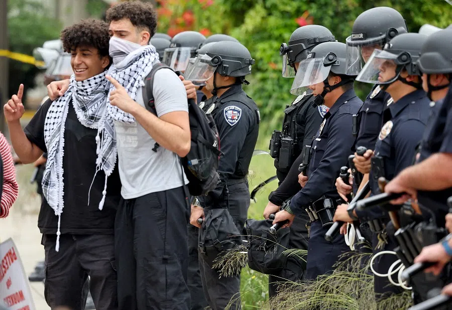 People wearing keffiyehs pose for a photo as LAPD officers keep watch after police cleared a new encampment of pro-Palestinian protestors on the UCLA campus on 23 May 2024 in Los Angeles, California.  (Mario Tama/Getty Images via AFP)