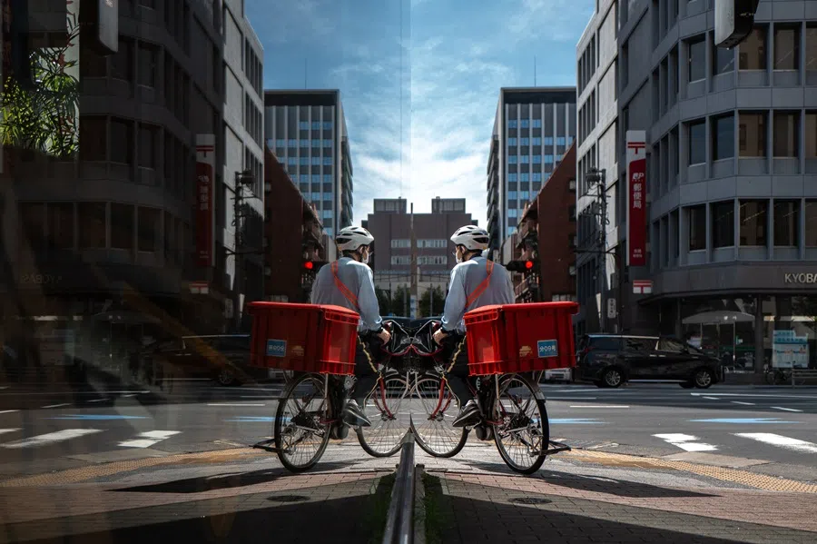 A postal worker is seen reflected in glass while riding a bicycle in the Ginza district of Tokyo on 8 October 2025. (Philip Fong/AFP)