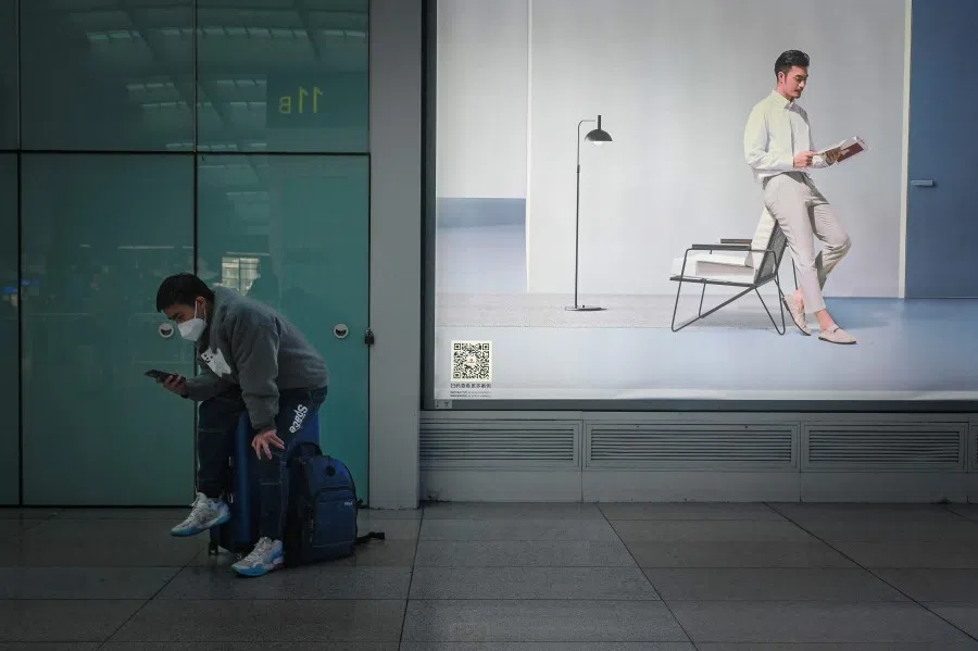 A man sitting on his luggage uses his mobile phone in front of an advertisement at Beijing south railway station in Beijing on 8 December 2022. (Wang Zhao/AFP)