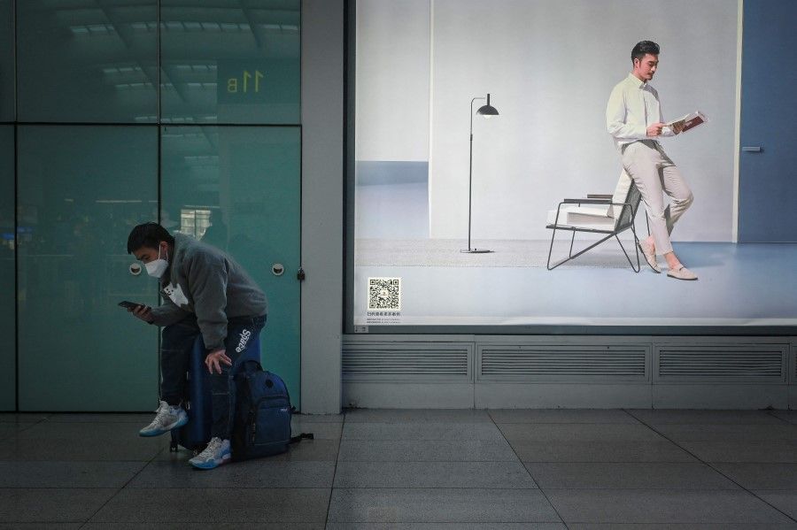 A man sitting on his luggage uses his mobile phone in front of an advertisement at Beijing south railway station in Beijing on 8 December 2022. (Wang Zhao/AFP)