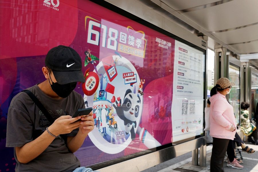 A man, wearing a face mask following the Covid-19 outbreak, uses his mobile phone in front of a Unionpay advertisement for the 618 shopping festival displayed at a bus stop, in Beijing, China, 14 June 2022. (Carlos Garcia Rawlins/Reuters)