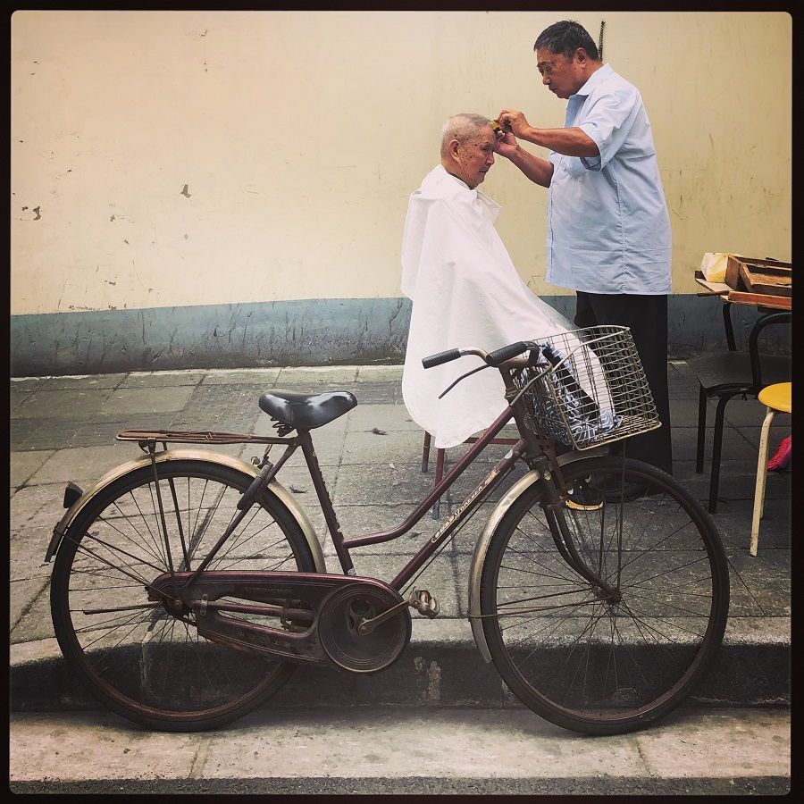 This barber, well dressed for good measure to attract customers, applies his grooming techniques to one resident in his territory on a city sidewalk in Huangpu, Shanghai.