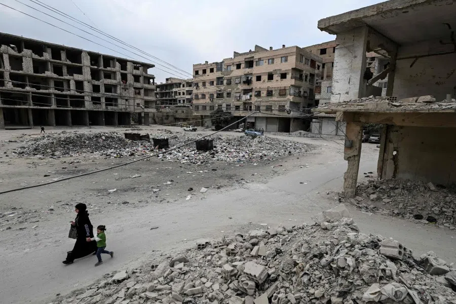 A woman and a child walk beneath electrical cables strung between damaged buildings in the Ain Tarma area, on the eastern outskirts of the capital Damascus, on 4 November 2025.