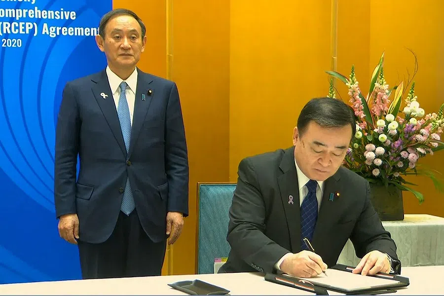 A screen grab taken from Vietnam Host Broadcaster's 15 November 2020 live video shows Japan Prime Minister Yoshihide Suga (left) standing next to Japan Minister of Economy, Trade and Industry Hiroshi Kajiyama as he signs the agreement during the signing ceremony for the RCEP trade pact at the ASEAN summit that is being held online in Hanoi. (Handout/Vietnam Host Broadcaster/AFP)