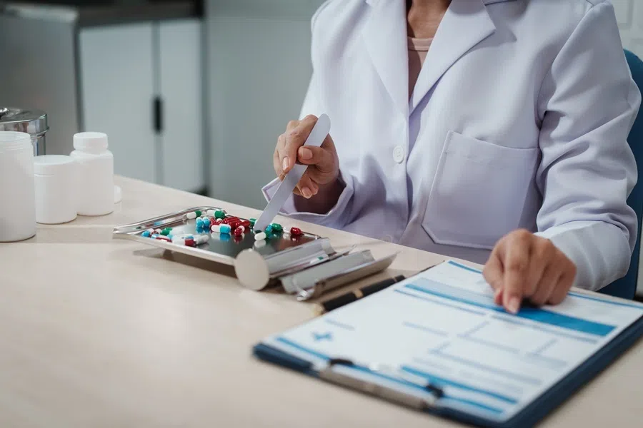 Pharmacist in white coat dispensing colorful pills with spatula onto tray beside prescription form on clipboard in pharmacy lab, close-up hands and medicine bottles.
