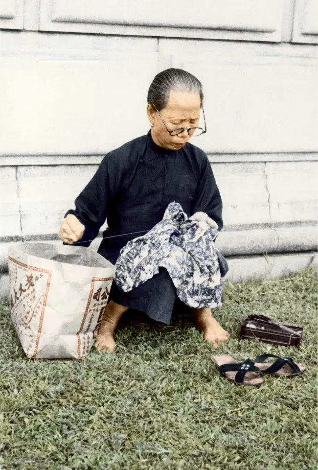 In the 1950s, an elderly woman mends clothes on the street to earn a small income to support her family.