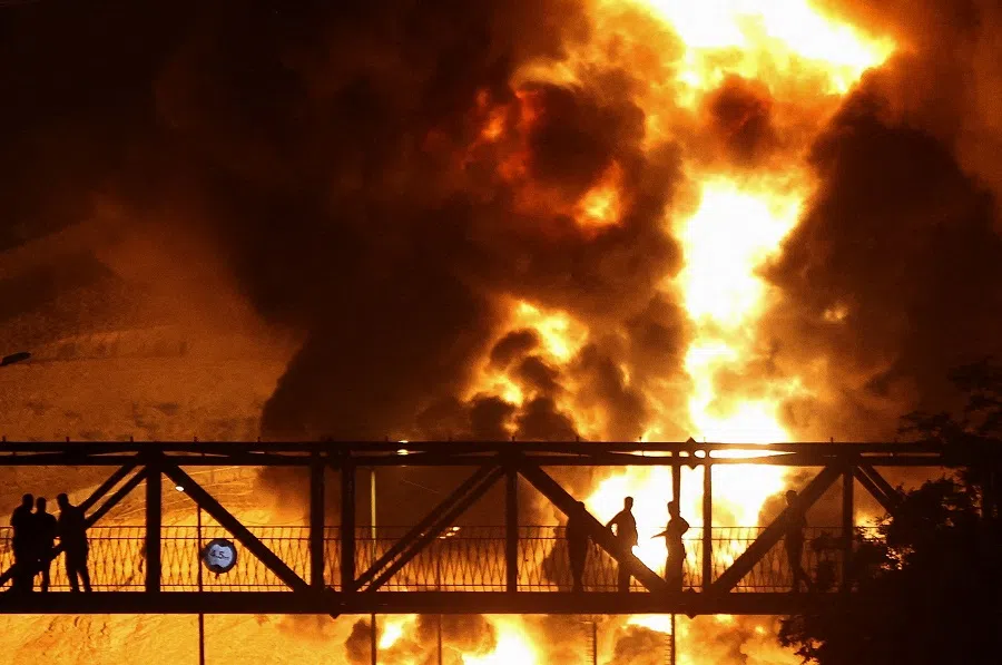 People watch from a bridge as flames from an Israeli attack rise from Sharan Oil depot, following Israeli strikes on Iran, in Tehran, Iran, on 15 June 2025. (Majid Asgaripour/West Asia News Agency via Reuters)
