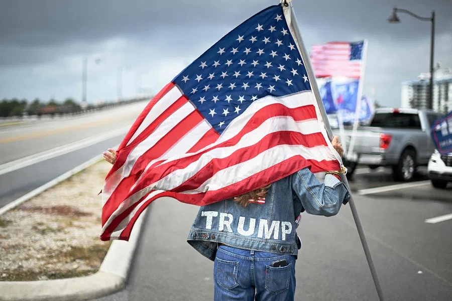 A supporter of US President-elect Donald Trump carries a US flag as she and others wait to see the Trump motorcade drive by on his way to the airport from Mar-a-Lago, in West Palm Beach, Florida, US, on 14 December 2024. (Roberto Schmidt/AFP)