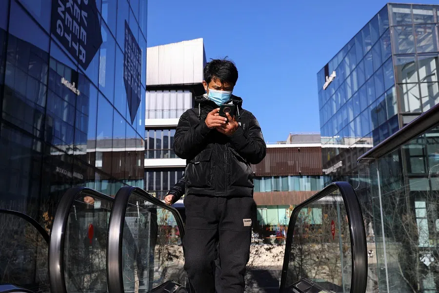 A man wearing a mask checks his phone as he rides on an escalator at a shopping complex in Beijing, China, 1 December 2021. (Tingshu Wang/Reuters)