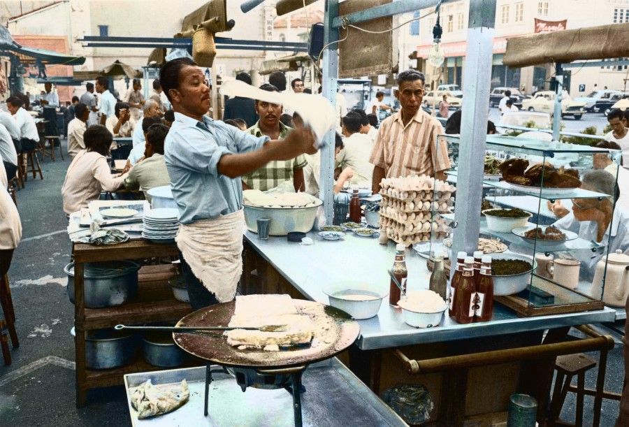 A roti prata stall in a market, 1960s. A worker is flipping a prata, something unique to Indian cuisine.