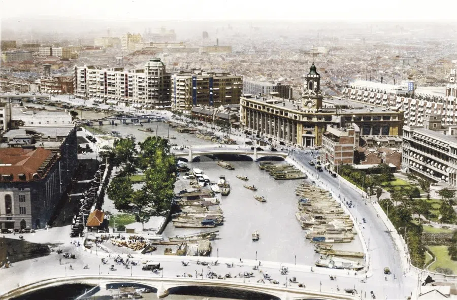 A view of the Sichuan Road Bridge and Shanghai Post Office headquarters during the days of the Republic. Boats sail under the bridge - sampans, sailboats, and Western-style ferries ply the Suzhou Creek, also called the Wusong River - while bicycles, cars, and traditional rickshaws mill around the bridge and street.