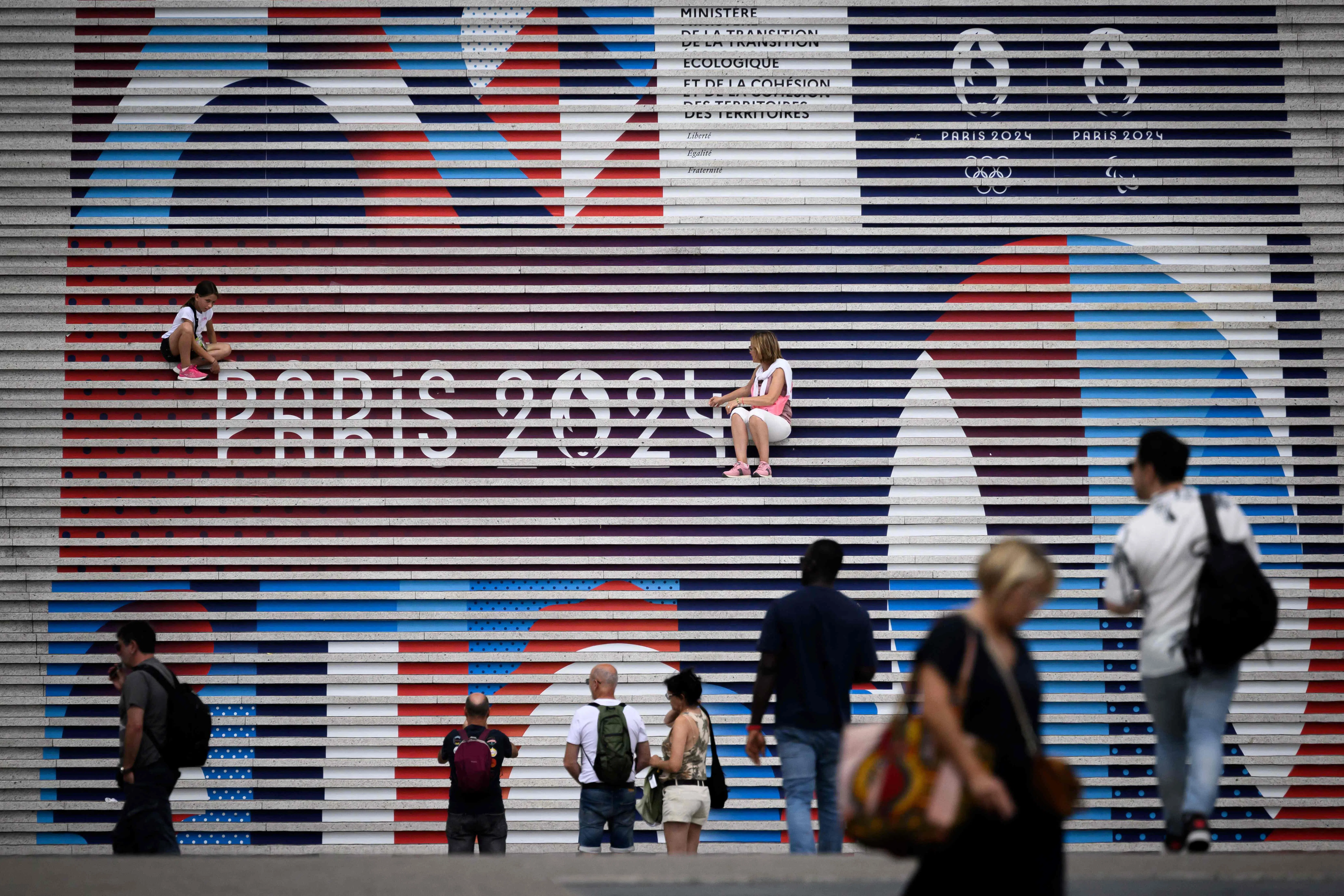 People pose on stairs near the logo of Paris 2024 Olympics Olympic games a few days before the opening ceremony, at La Defense business district, West of Paris, on 22 July 2024.