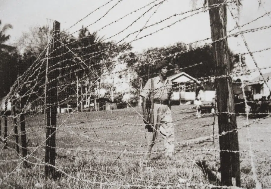 A photo of an image showing barbed wire fences and police guard in a new village in Malaysia, from the book Moving Mountains: A Pictorial History of the Chinese in Selangor and Kuala Lumpur.