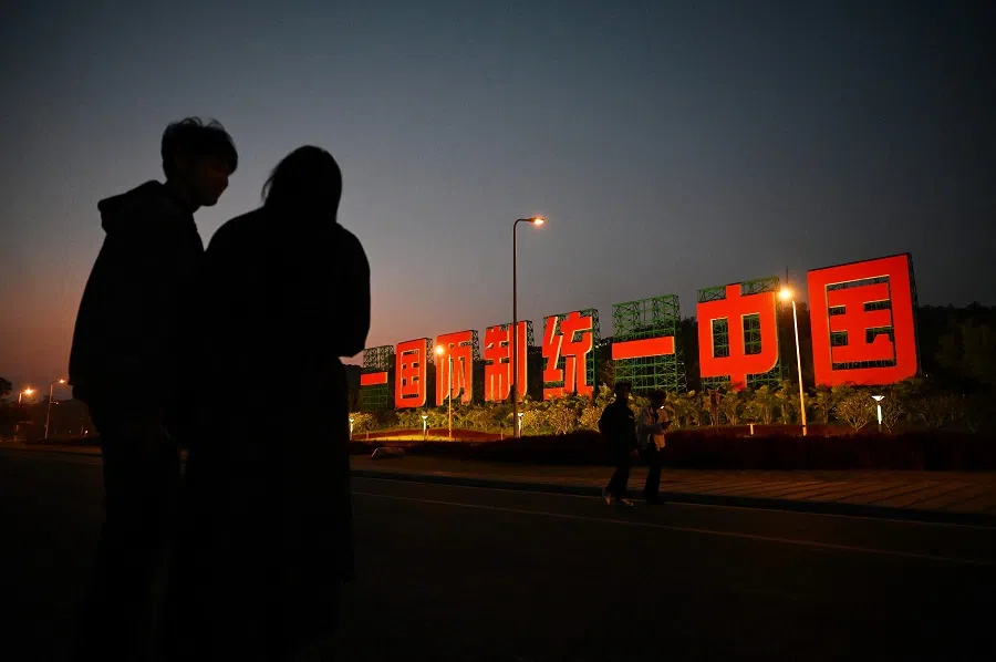 People stand near a giant propaganda slogan which reads "One Country, Two Systems, Reunify China", which can be seen from Taiwan's Kinmen Island, on a sidewalk on the waterfront in Xiamen, Fujian province, China, on 10 January 2024. (Greg Baker/AFP)