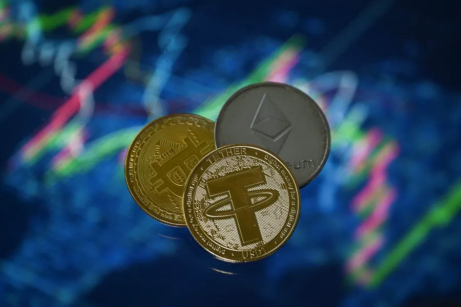 An illustration picture taken in London on 8 May 2022, shows gold-plated souvenir cryptocurrency Tether (USDT), Bitcoin and Etherium coins arranged beside a screen displaying a trading chart.