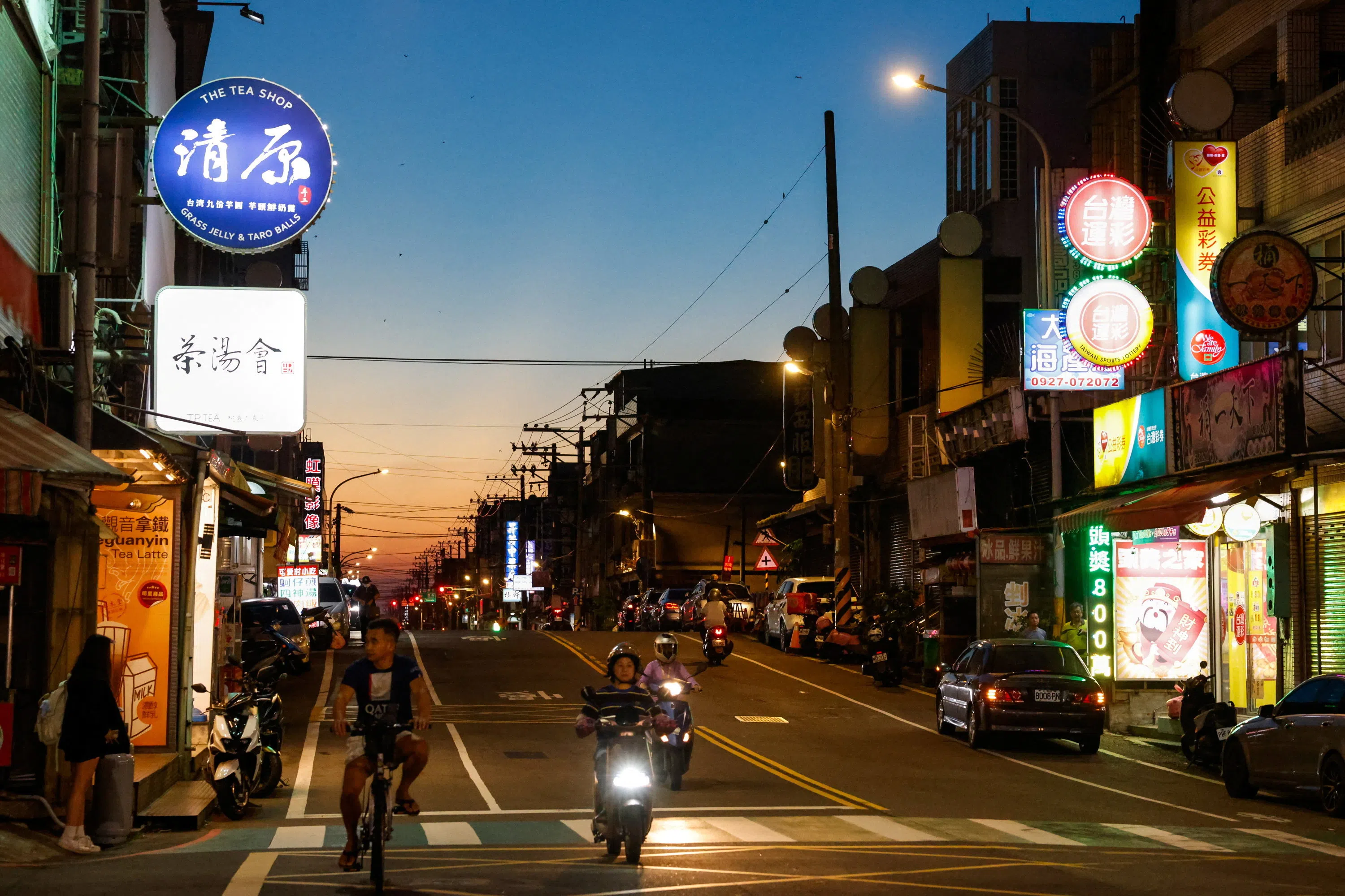 People travel along a street lined with shops and illuminated signs at dusk in Taoyuan, Taiwan, on 22 September 2026.