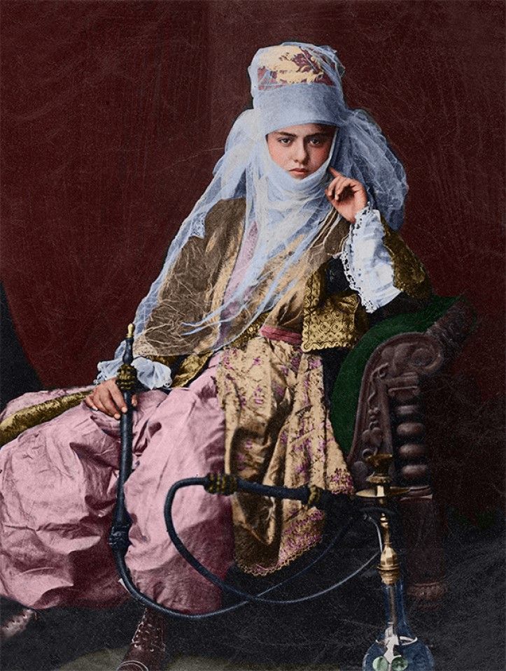 Turkey, 1880s. A high-born woman reclines on a sofa holding a nargile (also known as shisha/hookah) pipe, with its rubber tubing connecting the vessel and the mouthpiece. A thin veil covers her face and hat.