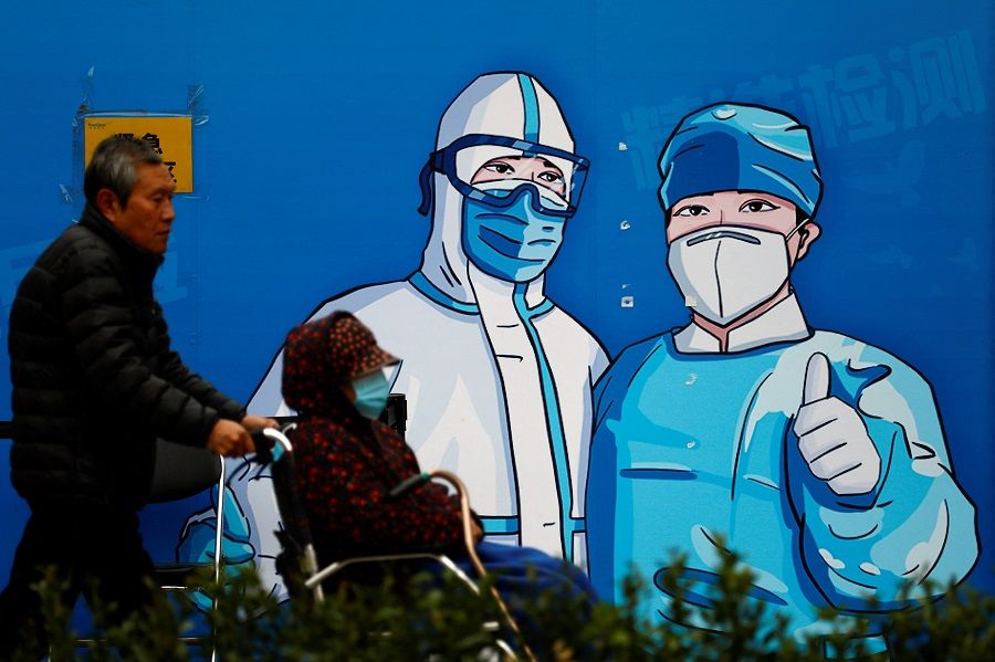 A man pushes a woman in wheelchair past a poster with a graphic of medical workers at a nucleic acid testing booth in Beijing, China, 27 October 2022. (Tingshu Wang/Reuters)